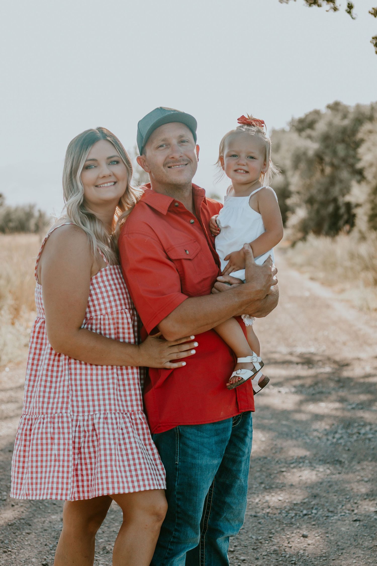 Happy family of three posing on a dirt path, parents in red outfits holding a toddler girl with a red bow.