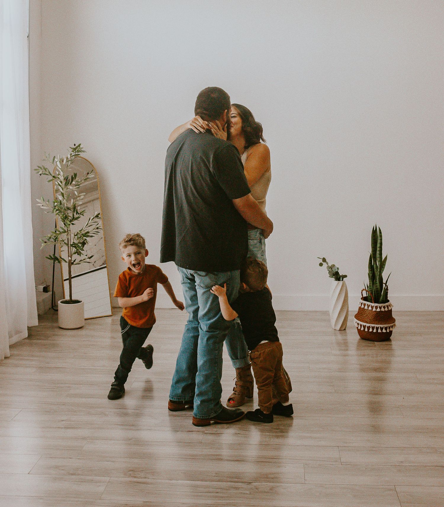 A family of four shares a tender moment in a bright room, with two children playing around their embracing parents.