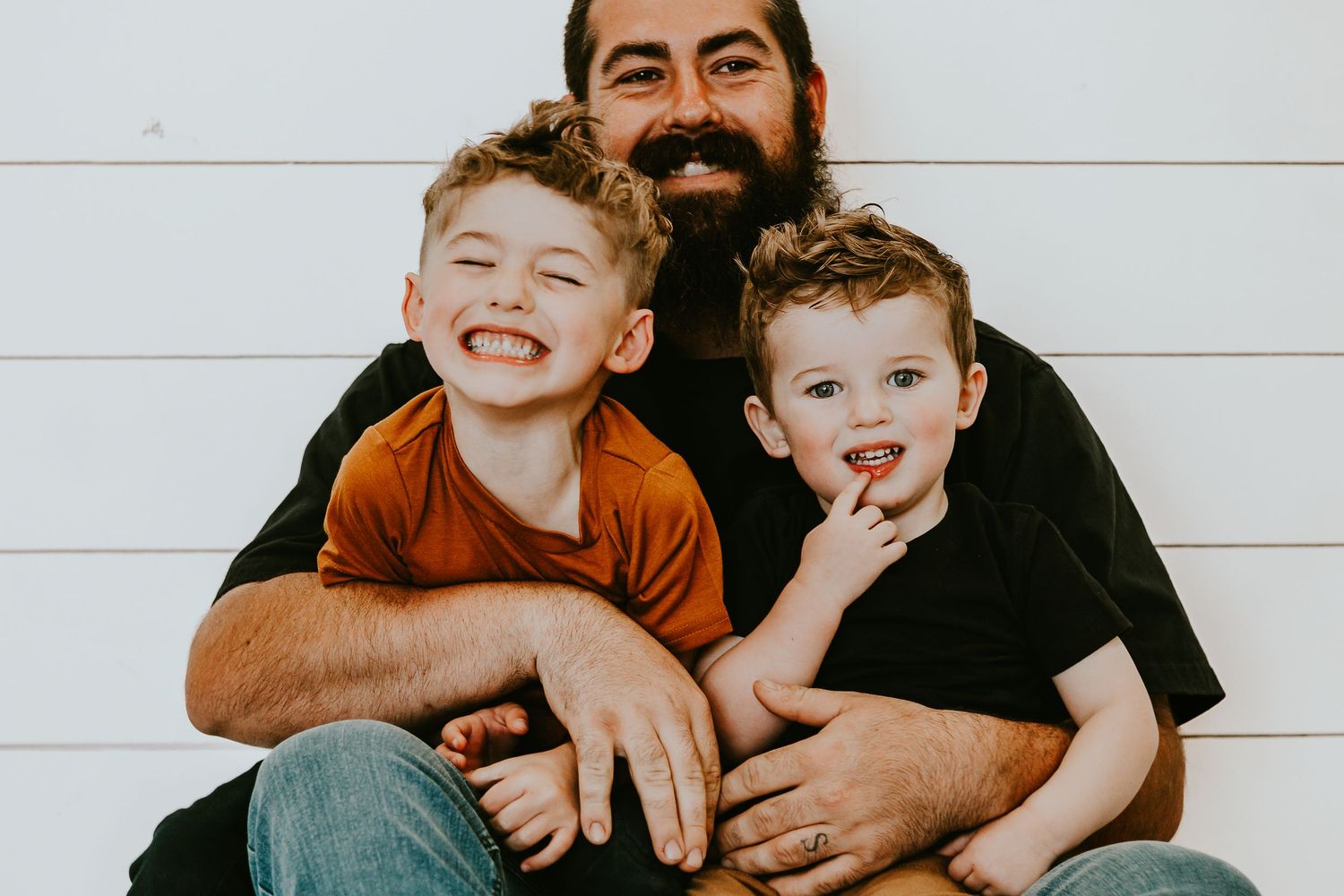 Bearded father smiling and hugging two young boys against a white wooden wall in a warm family portrait.