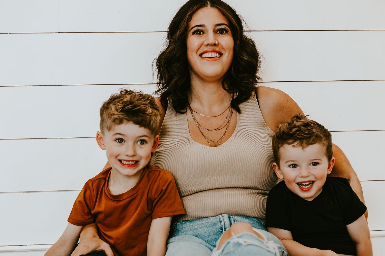 A smiling woman poses with two young boys in front of a white shiplap wall in a casual family portrait.