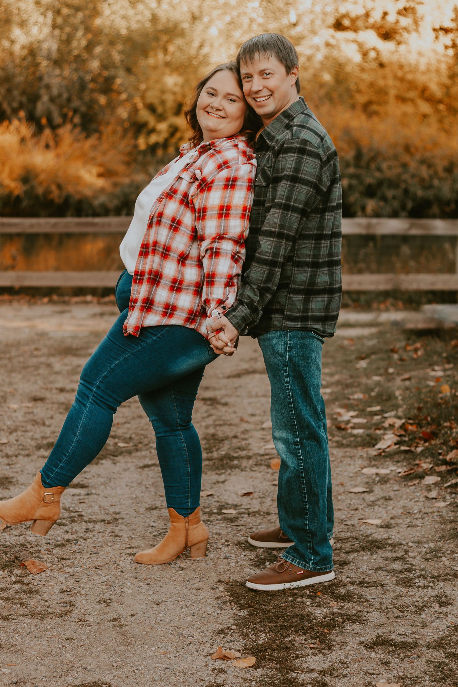 Couple holding hands in matching flannel shirts and jeans, smiling outdoors in autumn setting with golden foliage.