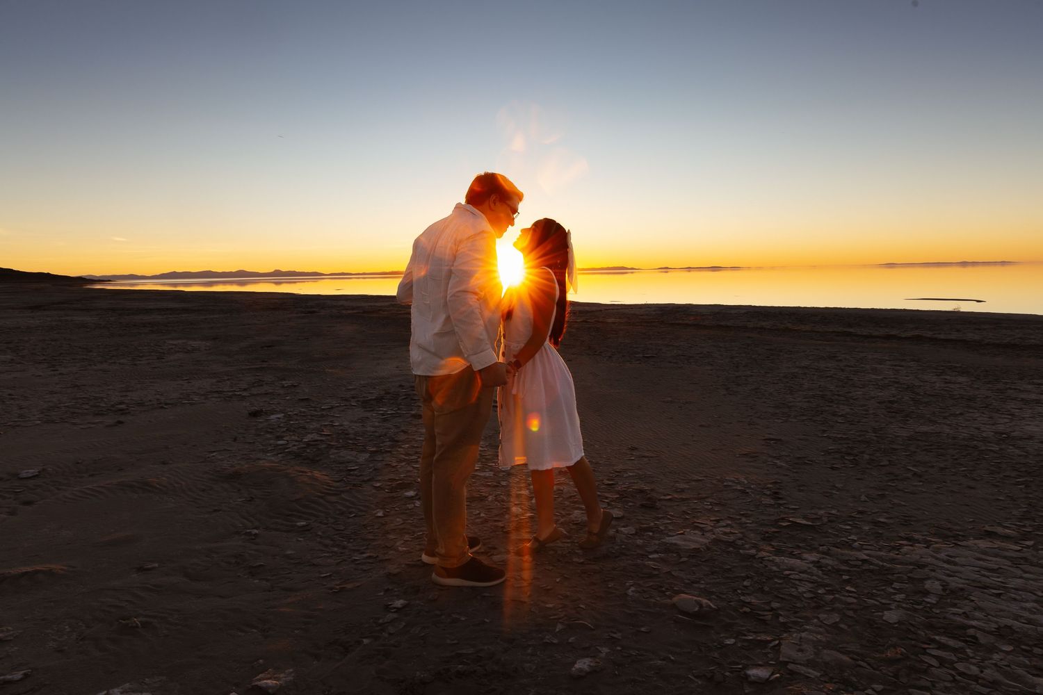 Couple sharing a romantic sunset kiss on a beach, silhouetted against a golden horizon over calm waters.