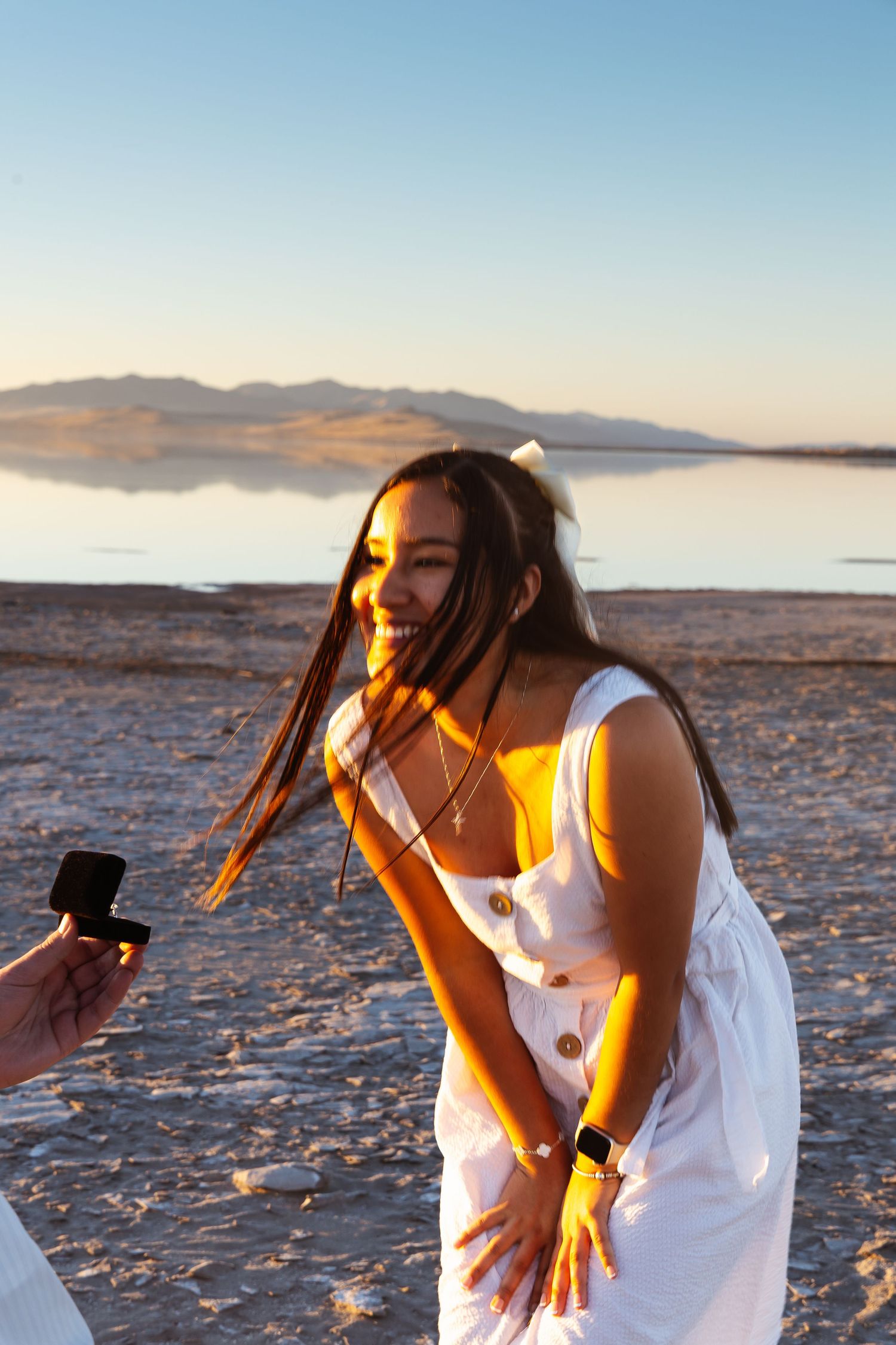 A joyful woman in a white dress reacts to a marriage proposal at sunset by a serene lake with mountains.