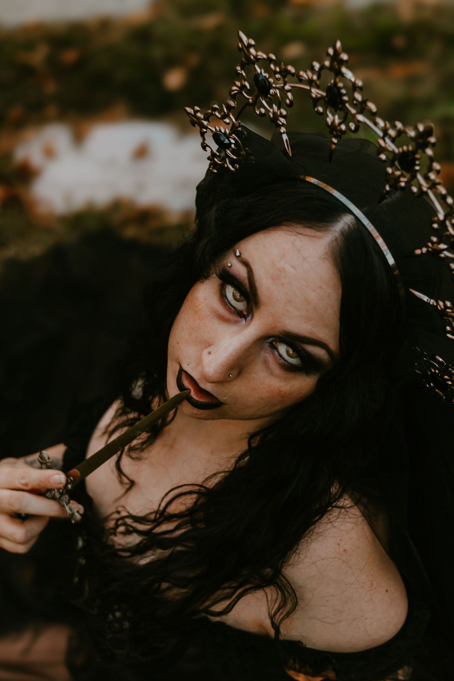 Gothic woman in dark crown and makeup holding a black rose, dramatic dark fantasy portrait outdoors.