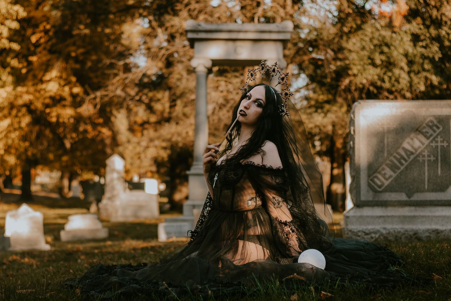 Woman in black gothic gown and crown poses dramatically among cemetery headstones with autumn foliage backdrop.