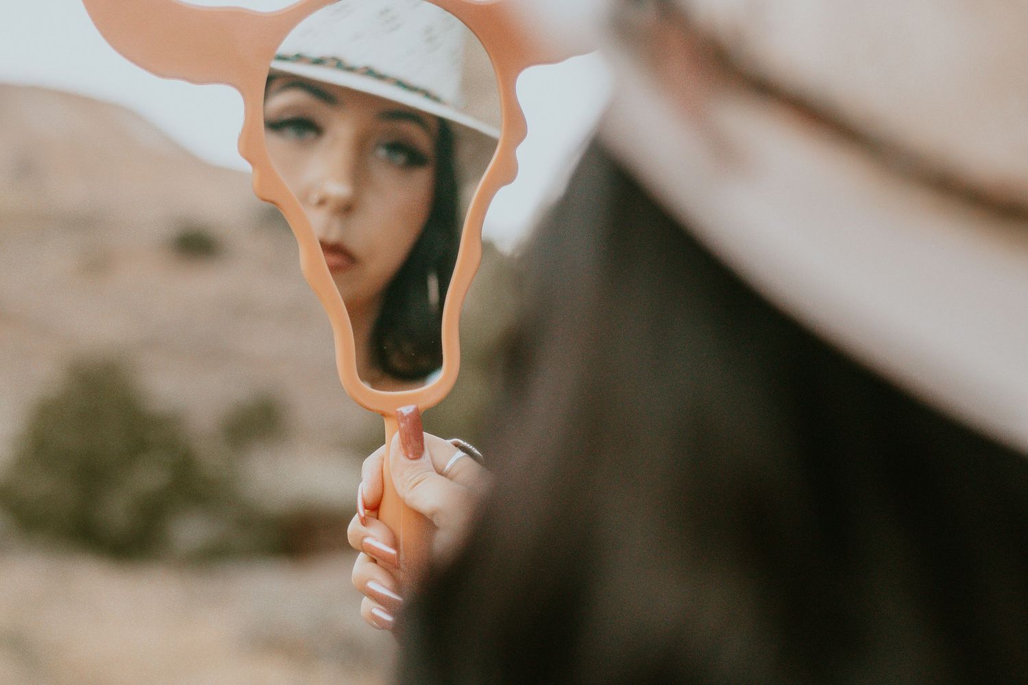 A young woman with dark hair holds a decorative hand mirror reflecting her face outdoors in a desert setting.