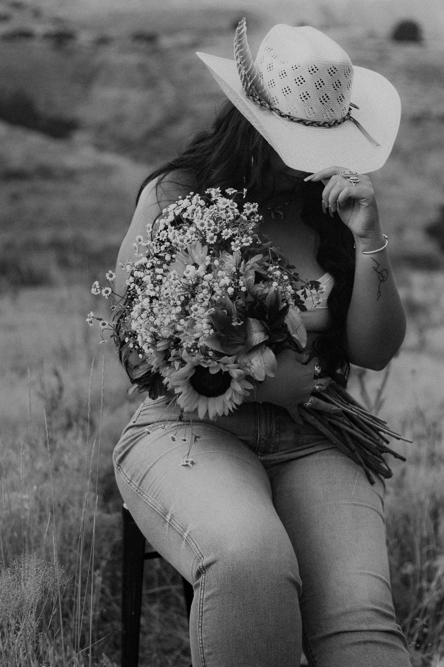 Black and white photo of a woman in a cowboy hat holding a wildflower bouquet with sunflowers while sitting outdoors.