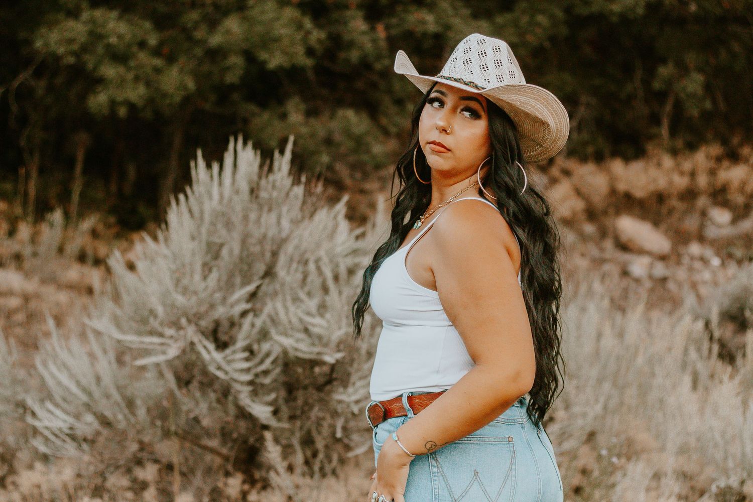 Woman in white crop top and cowboy hat posing outdoors in a desert landscape with sagebrush.