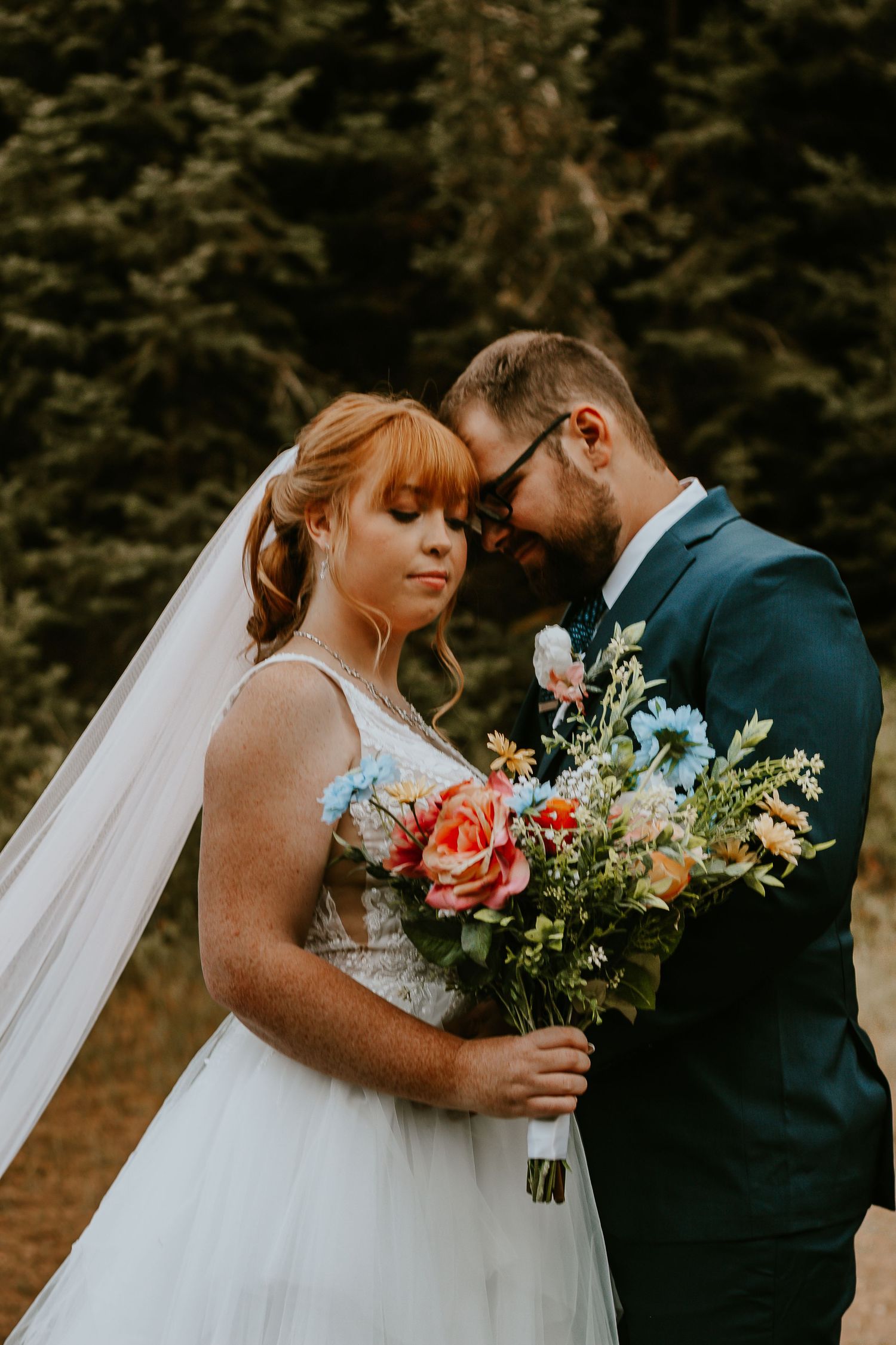 Bride and groom share an intimate forehead touch, bride holding a colorful wildflower bouquet in a lush outdoor setting.