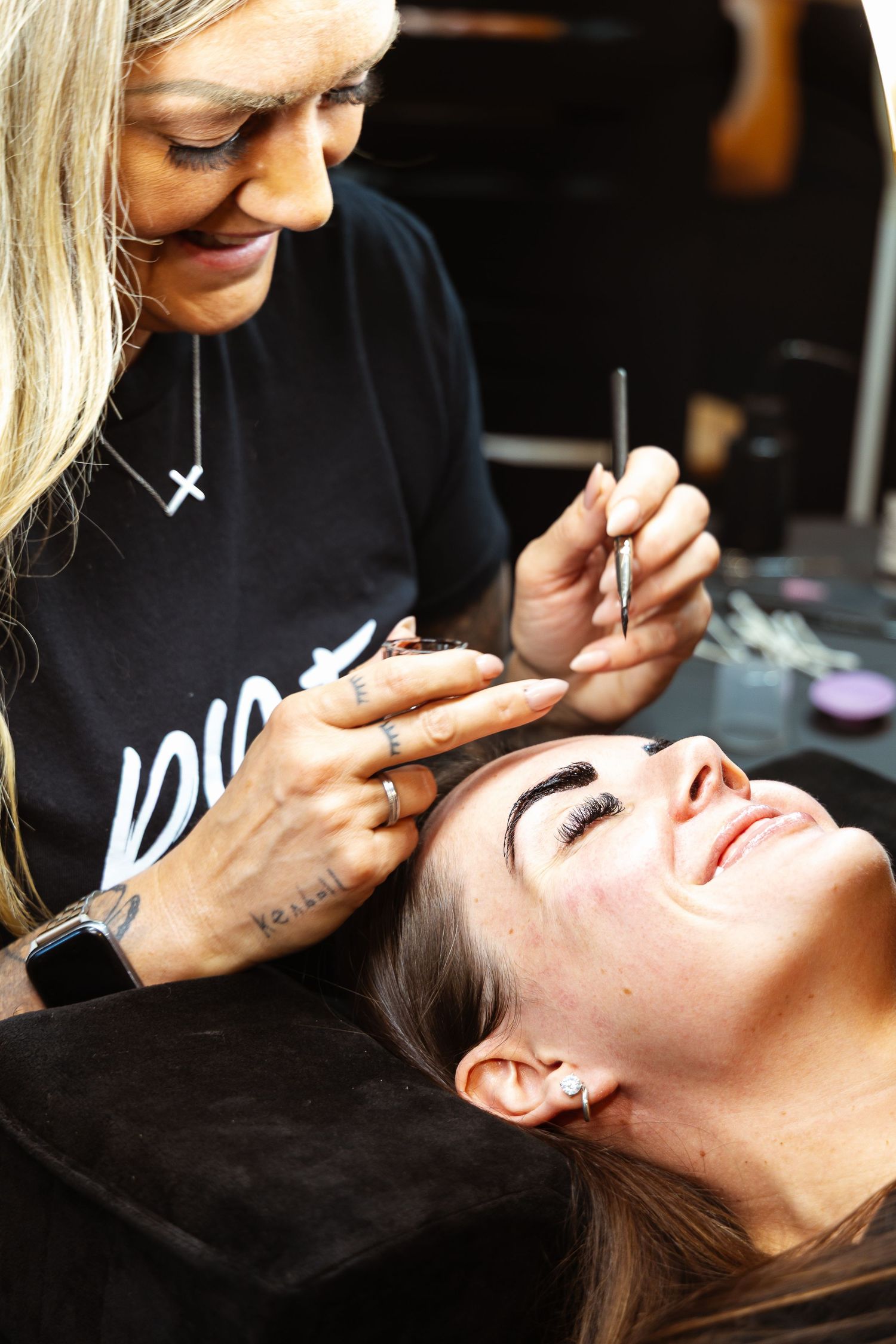 A makeup artist applies eyebrow makeup to a smiling woman lying back in a salon chair.