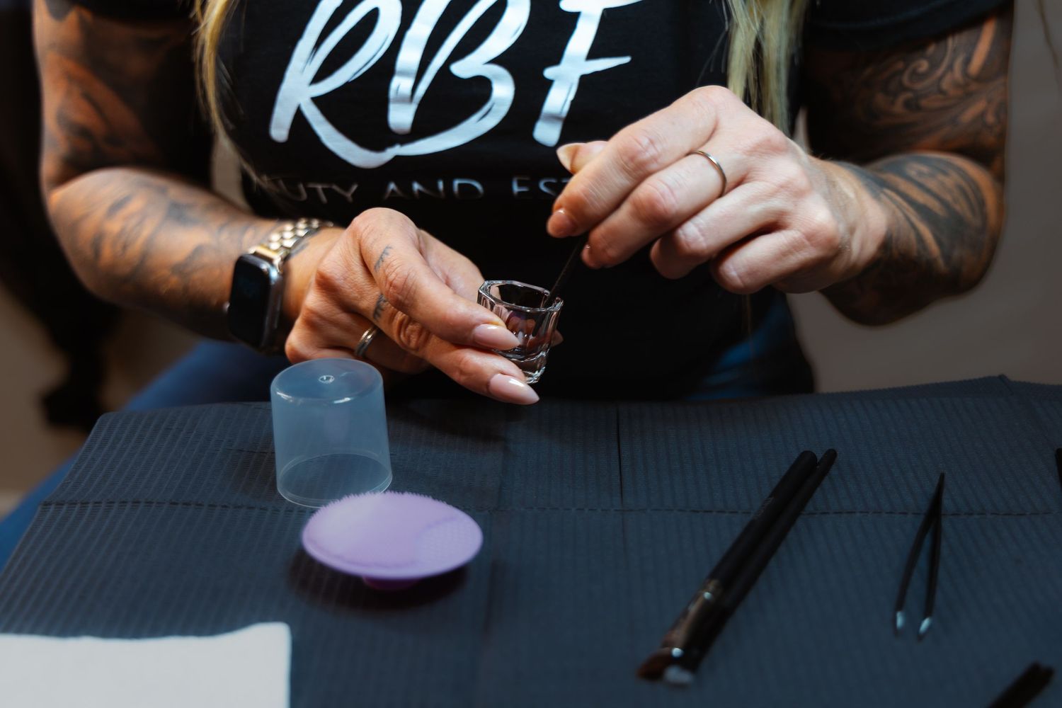 Tattooed woman in RBF shirt holding a shot glass over a black table with beauty tools and containers.