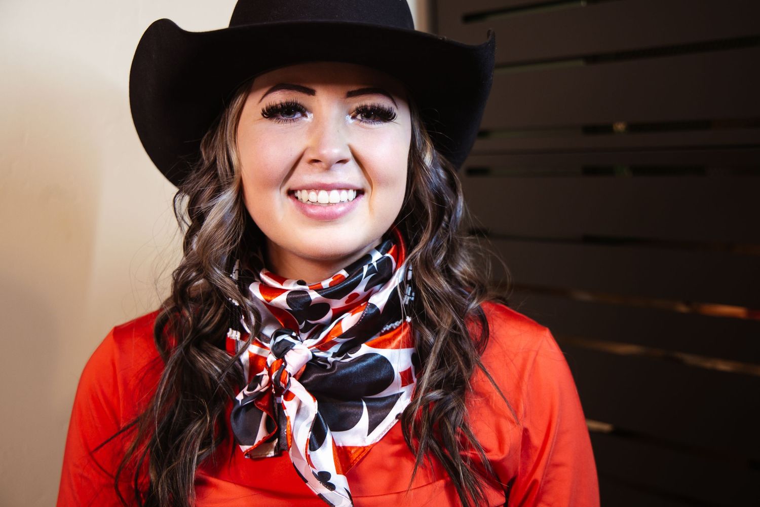 Smiling young woman wearing a black cowboy hat, red top, and colorful bandana scarf, posing indoors.