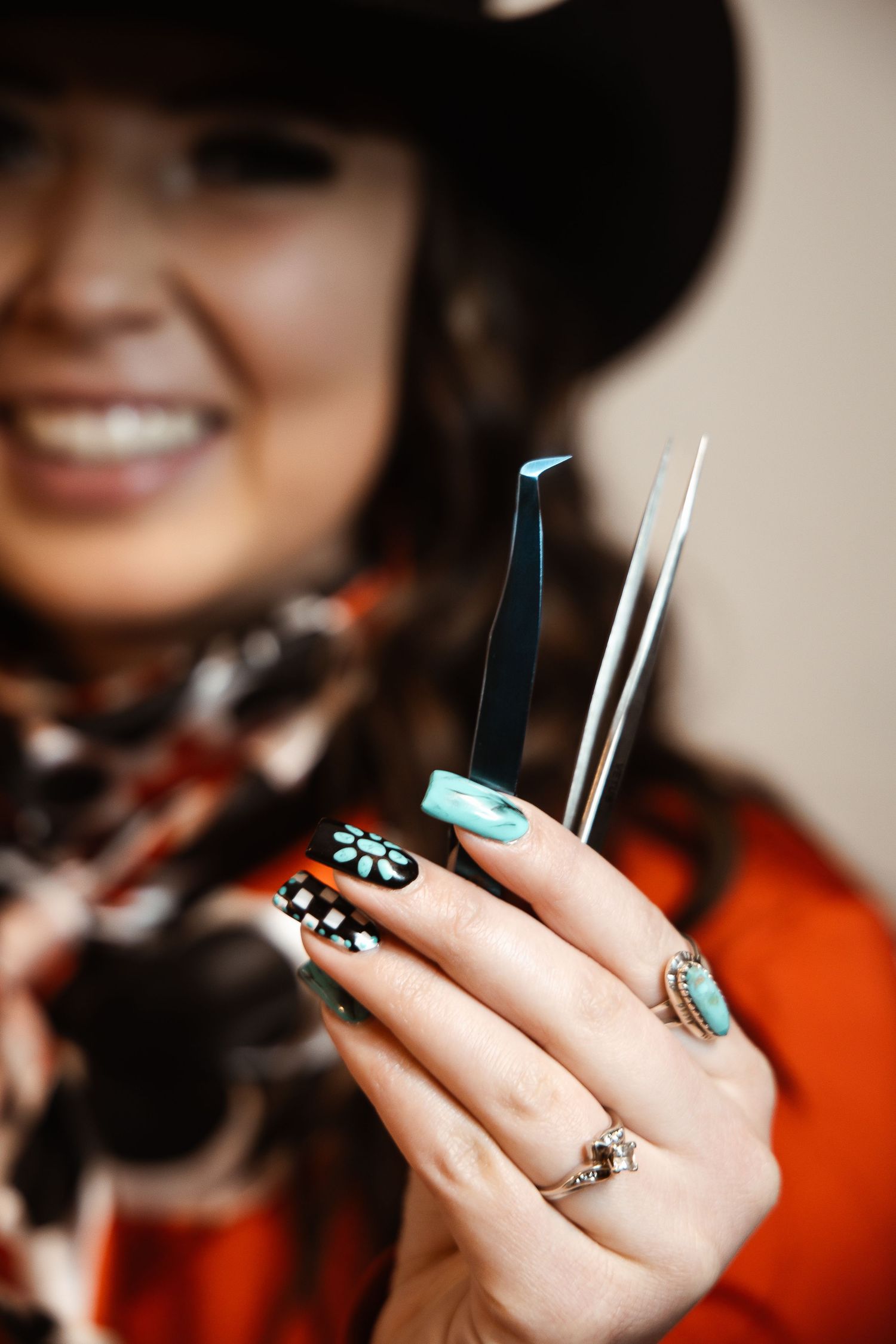 Woman in cowboy hat displaying intricate turquoise and black nail art with floral designs, holding tweezers.