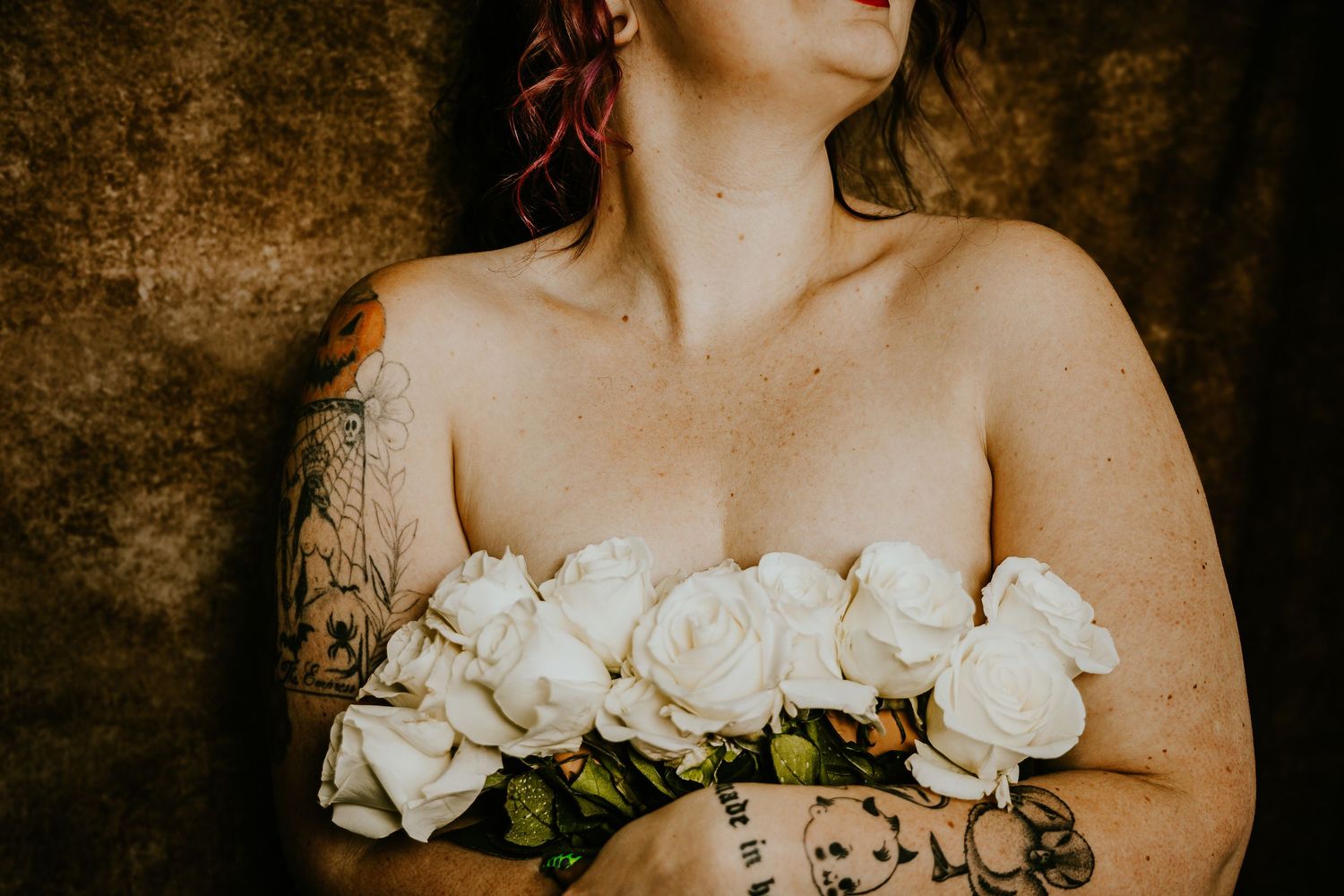 Tattooed woman holding white roses bouquet against rustic background in artistic portrait photography.