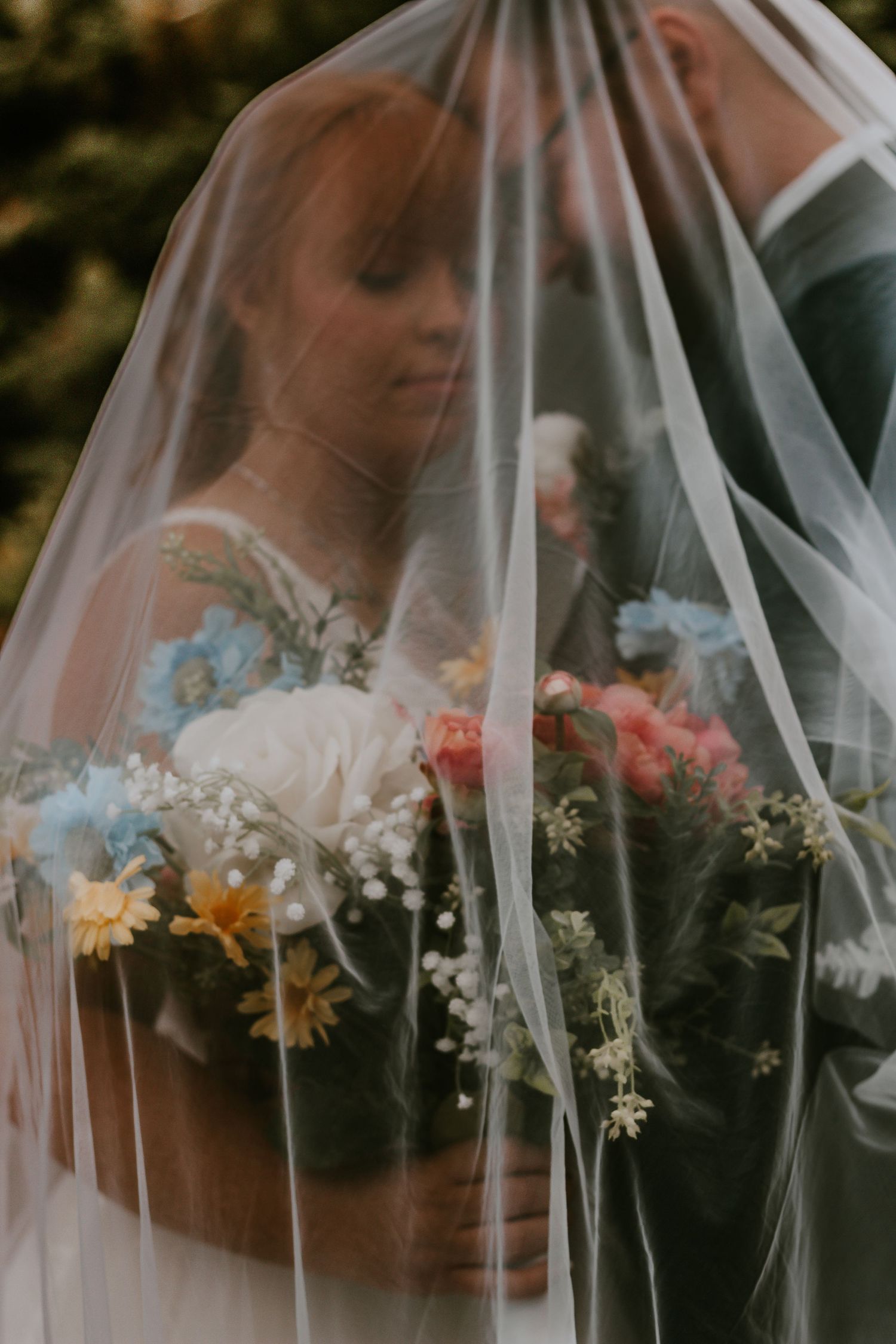 Bride and groom share a romantic moment under a sheer wedding veil, holding a colorful wildflower bouquet.