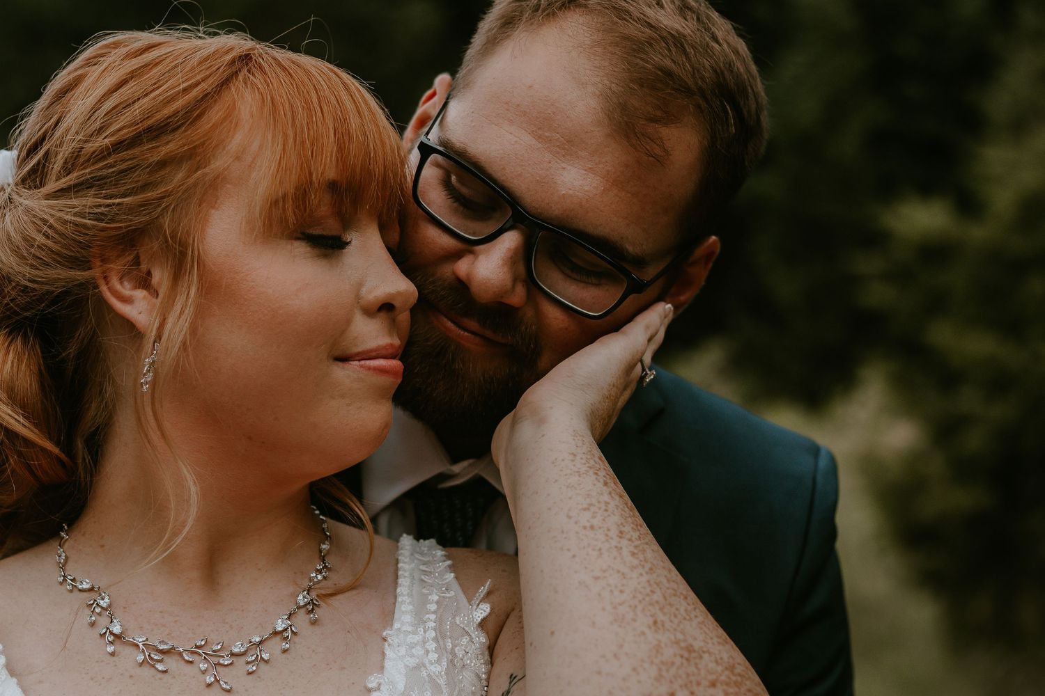 Romantic wedding couple closeup, bride in diamond necklace and groom in glasses sharing tender moment outdoors.