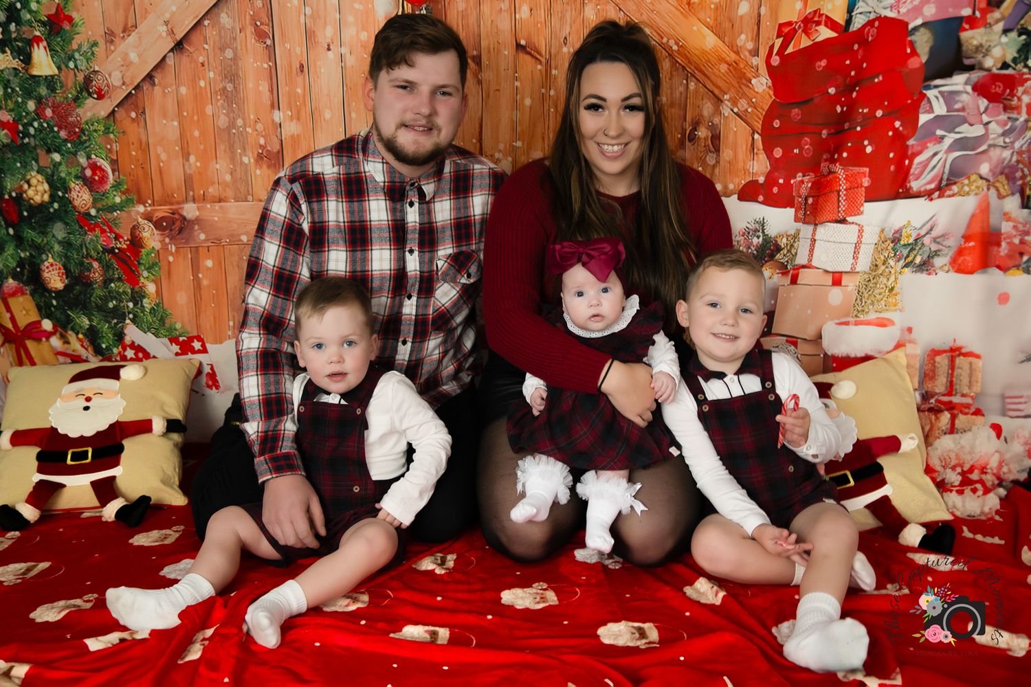 Family Christmas portrait with parents and three children on red blanket with decorated tree.