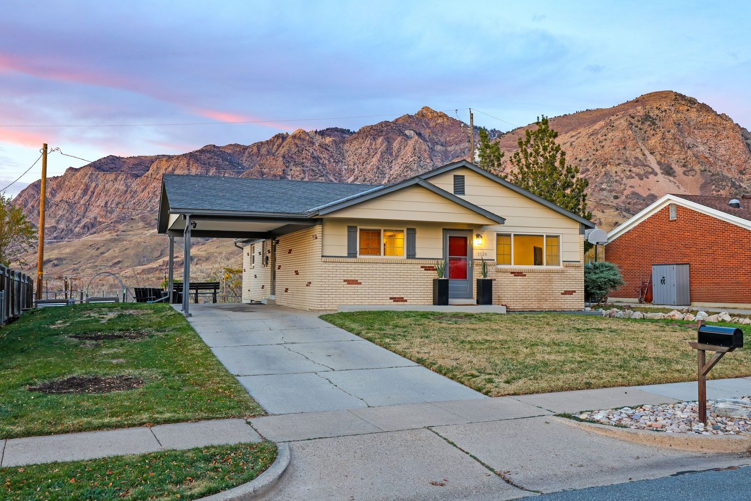 Ranch-style home with red door, carport, green lawn, and dramatic mountain backdrop at sunset in Utah. Captured by Slate Media (best in UT)