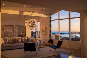 Modern living room with floor to ceiling windows offering stunning ocean views at sunset, featuring contemporary lighting and decor.