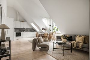 Modern Scandinavian open concept attic apartment with white kitchen, wood floors and grey sectional sofa in sunny setting.