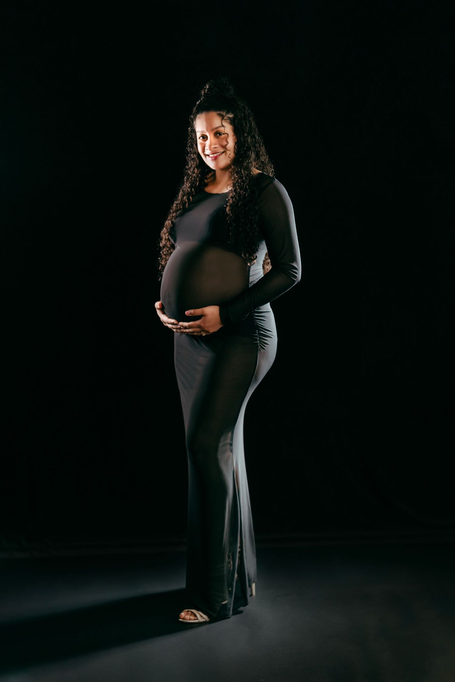 Maternity photoshoot series featuring a pregnant woman in a black dress against a dark background. Maternity Photographer
