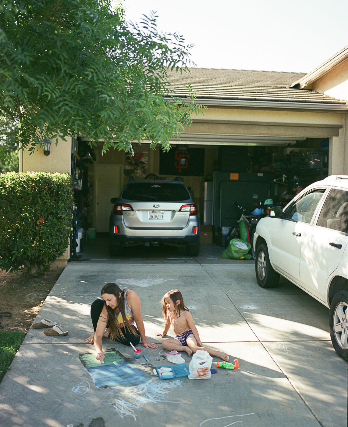 People drawing with chalk on a driveway next to parked cars on a sunny afternoon.