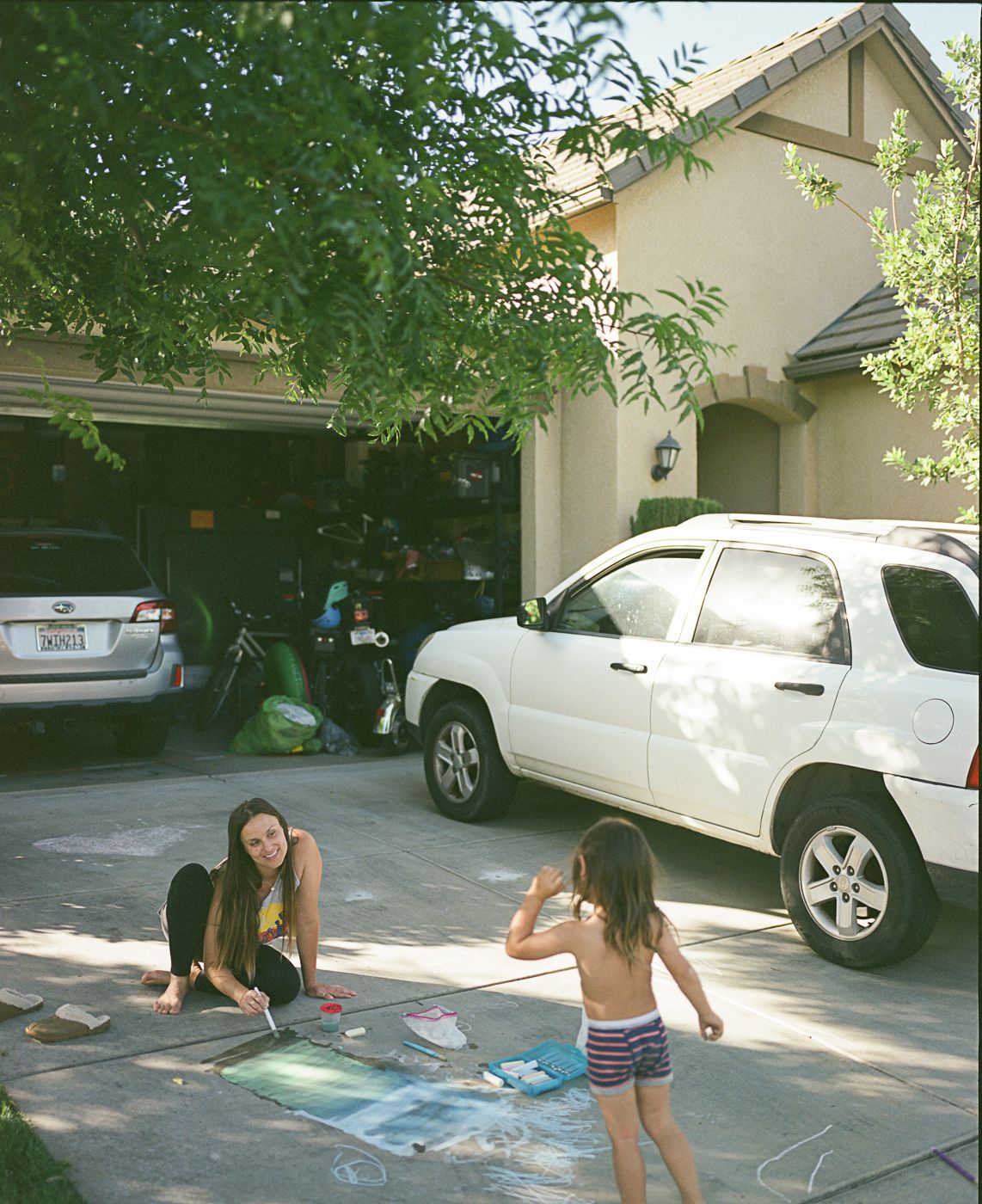 People drawing with chalk on a driveway with parked vehicles in the background.