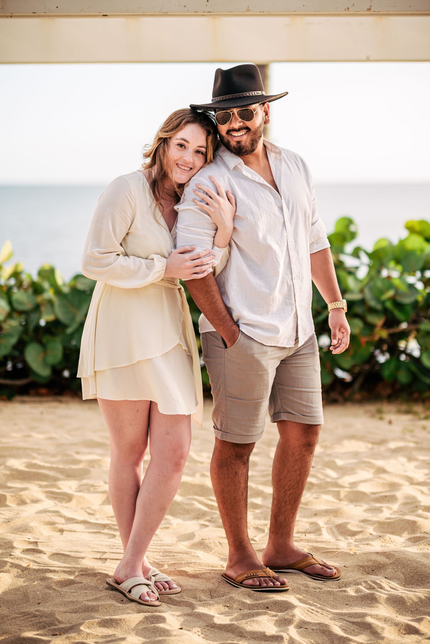 A casual couple poses on a sandy beach with ocean views in the background.