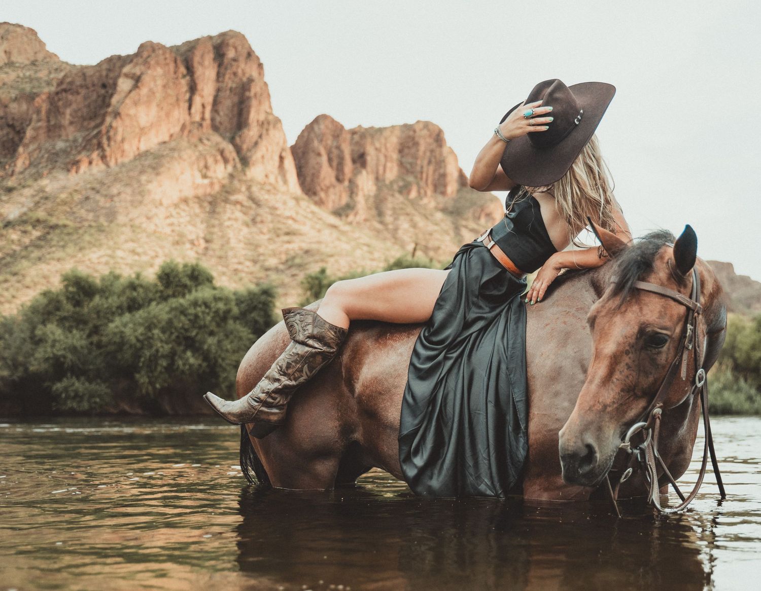 A rider in a flowing dress reclines on horseback in a scenic desert river with rugged mountains in the background.