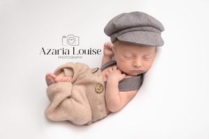 Sleeping newborn dressed in beige romper and gray newsboy cap rests peacefully on white background.