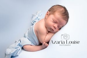A sleeping newborn wrapped in soft powder blue fabric rests peacefully against a light gray backdrop.