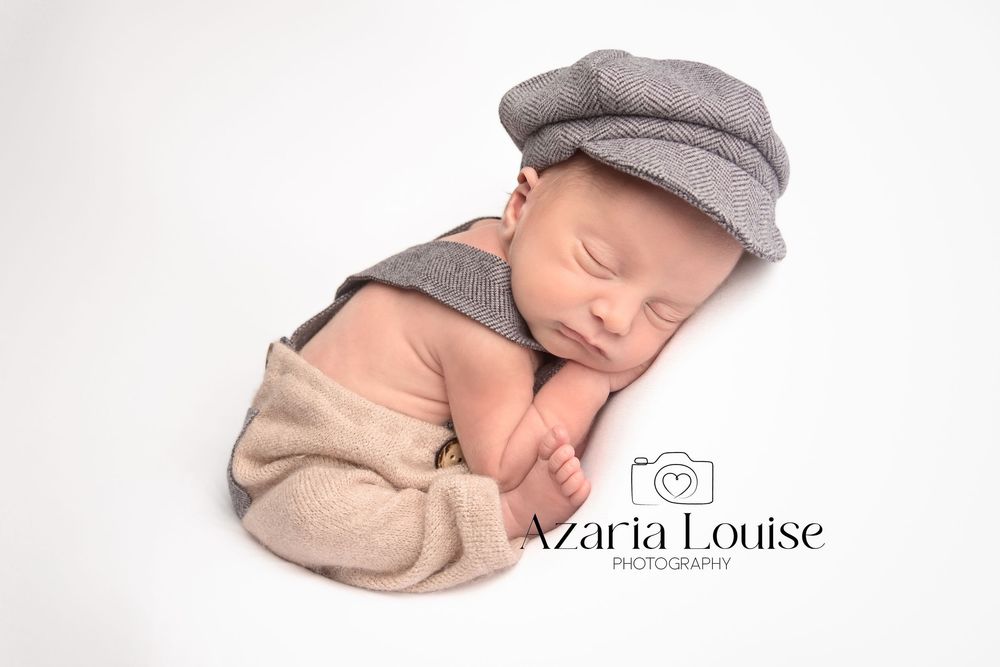 Sleeping newborn wearing a grey newsboy cap and beige wrap on white background during professional photography session.