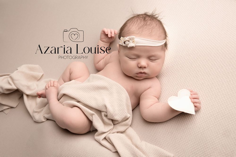 Newborn wearing delicate headband lying peacefully on beige backdrop with soft cloth and small paper heart.