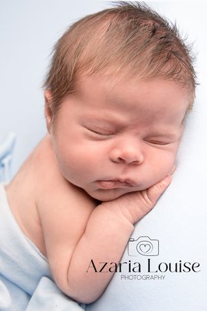 A peacefully sleeping newborn swaddled in light blue blanket rests with hand under cheek in professional portrait.