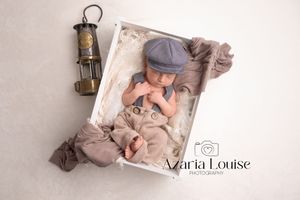 A newborn baby sleeps in a light gray outfit and cap next to a vintage lantern on a soft textured background.