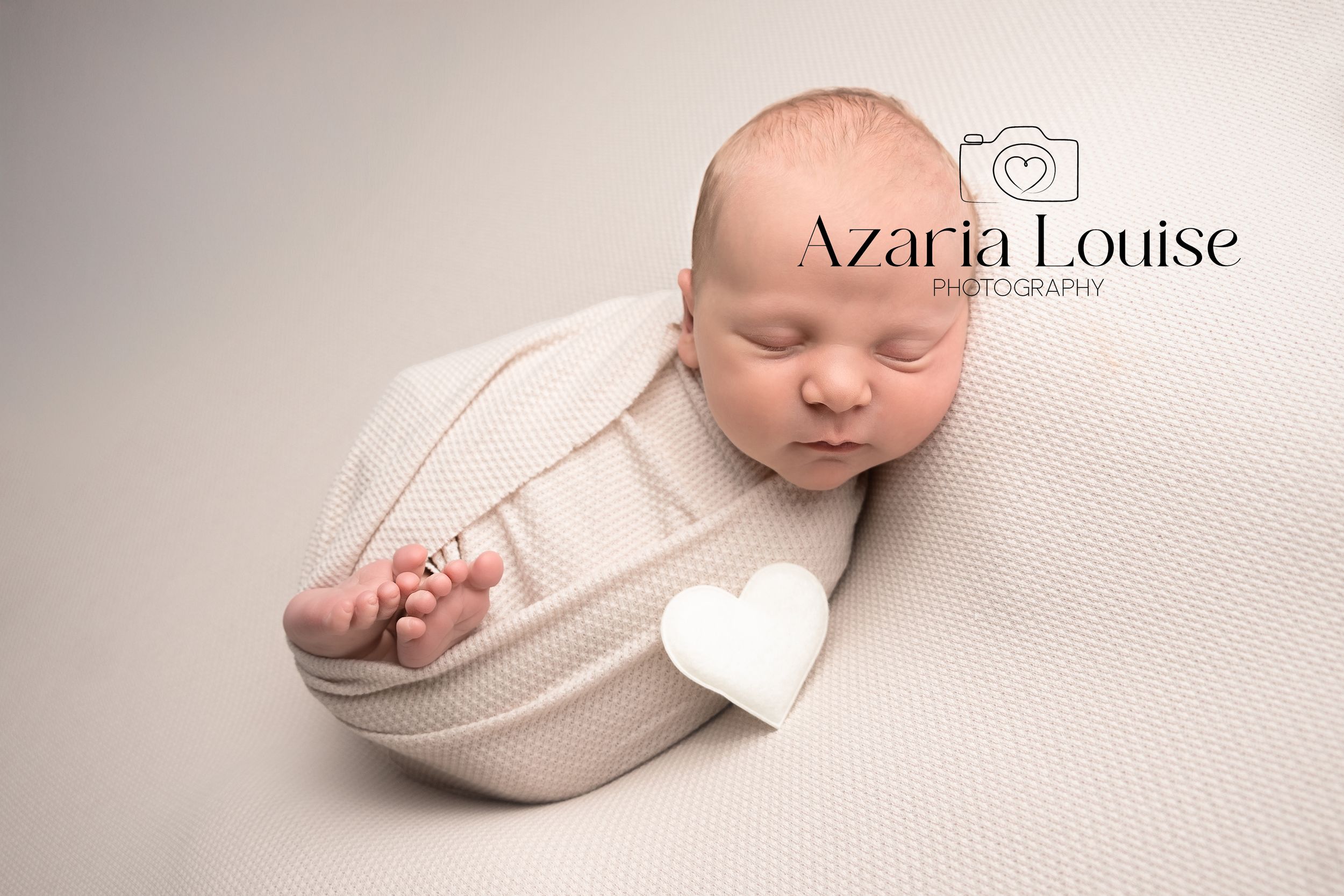 A peaceful newborn baby sleeps swaddled in soft white fabric on a cream-colored backdrop.