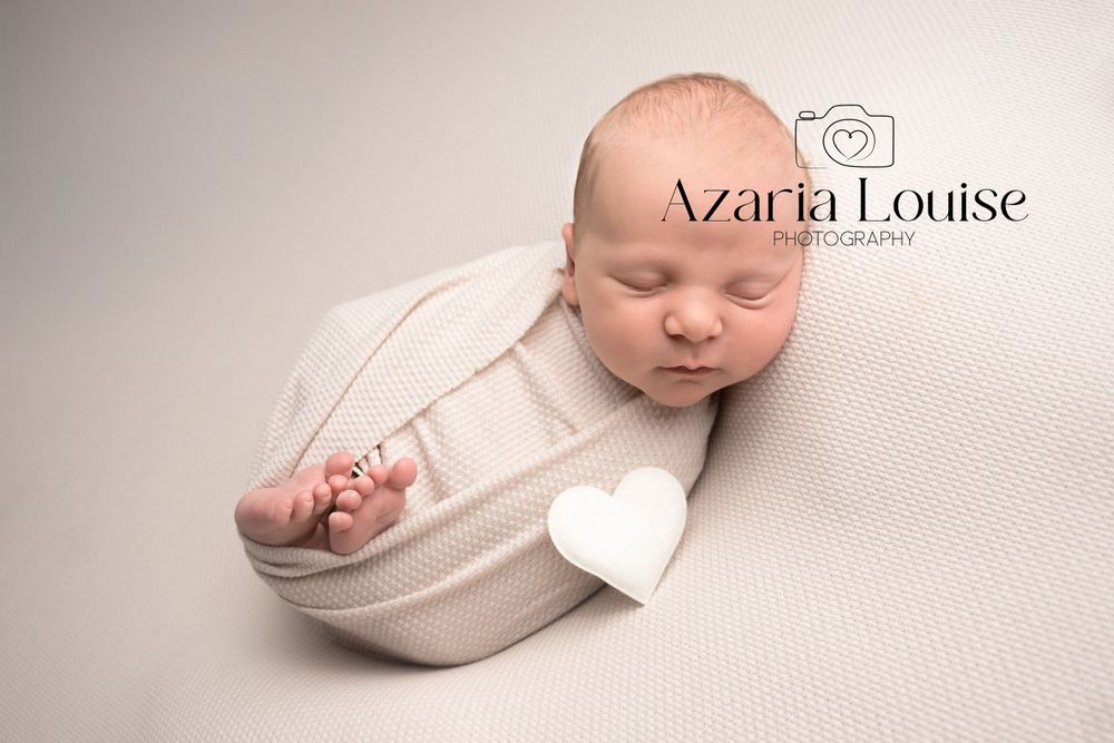 A peaceful newborn baby sleeps swaddled in soft white fabric on a cream-colored backdrop.