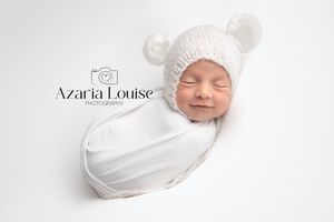 Sleeping newborn swaddled in white fabric wearing soft white bear ear hat during professional photo session.