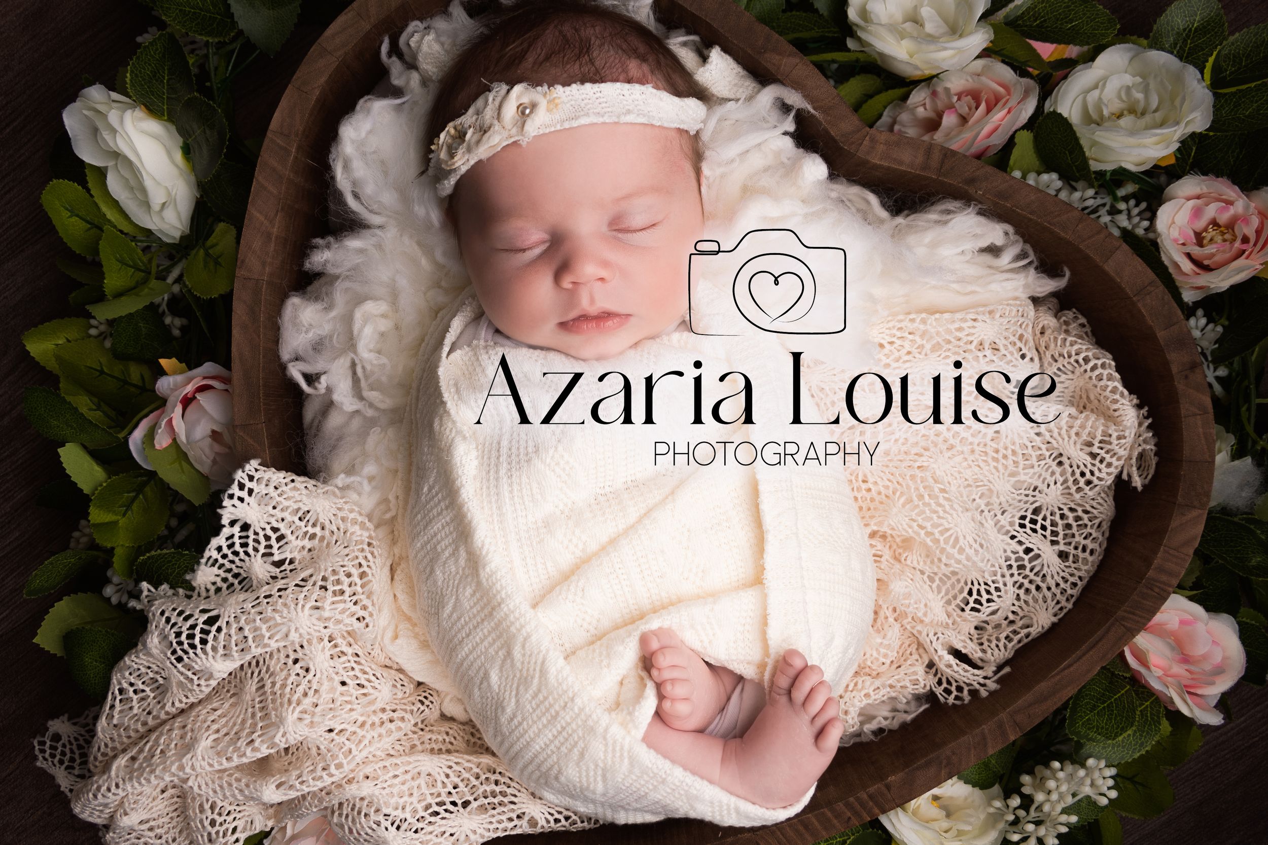 A sleeping newborn wrapped in delicate lace lies in a wooden heart bowl surrounded by soft pink roses.