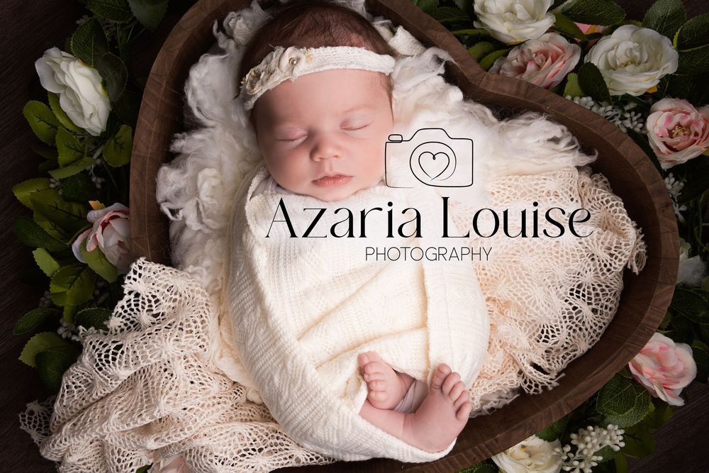 A sleeping newborn wrapped in delicate lace lies in a wooden heart bowl surrounded by soft pink roses.