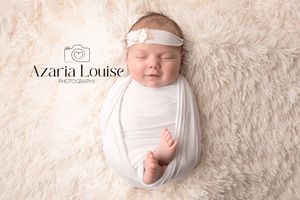A sleeping newborn wrapped in white fabric with a delicate headband lies on a soft cream-colored fur backdrop.