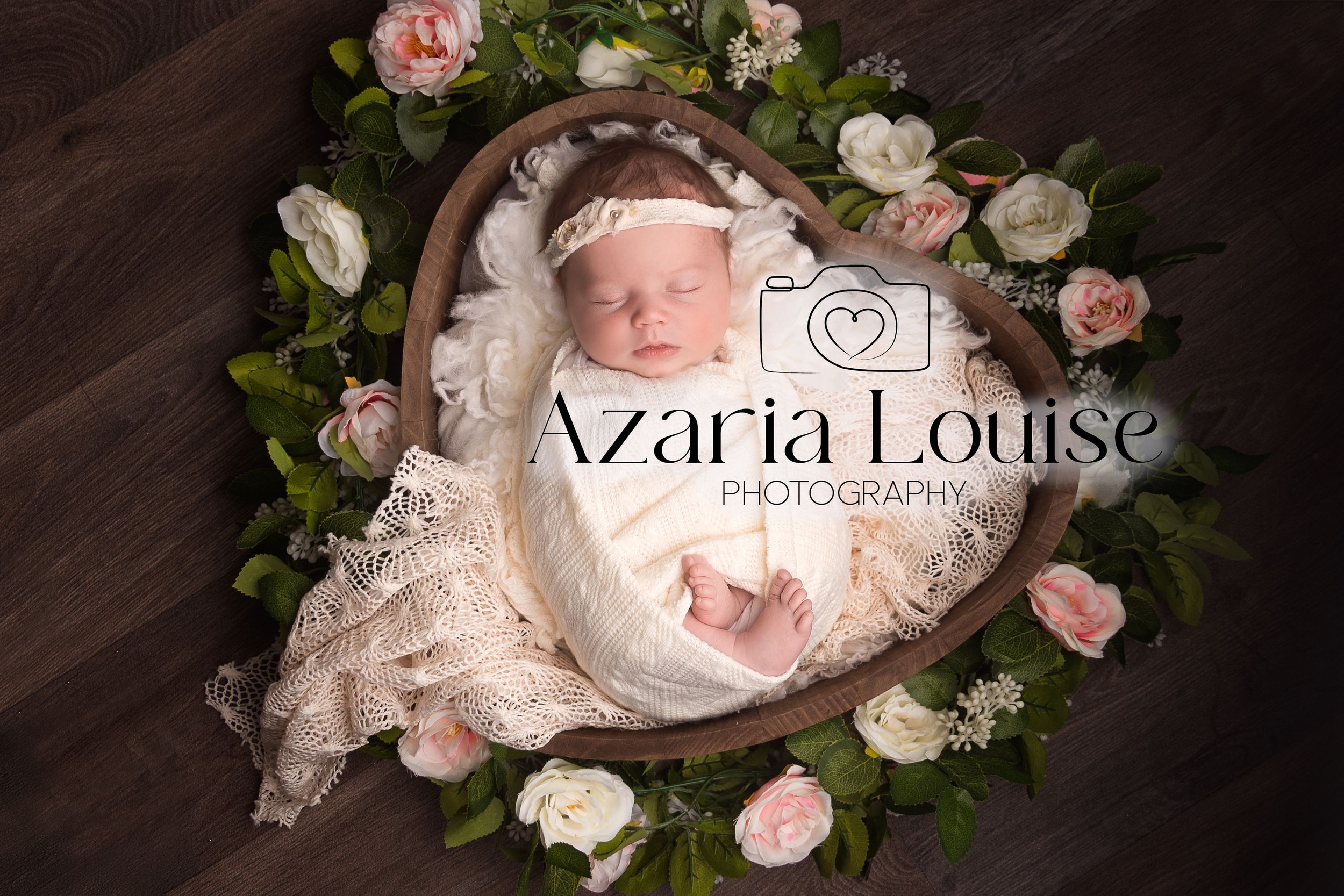 Newborn baby sleeping in a heart-shaped basket surrounded by pink and white roses and floral decor.