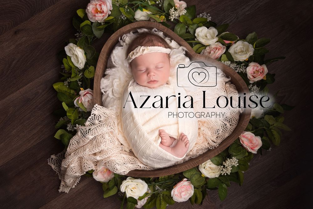 Newborn baby sleeping in a heart-shaped basket surrounded by pink and white roses and floral decor.