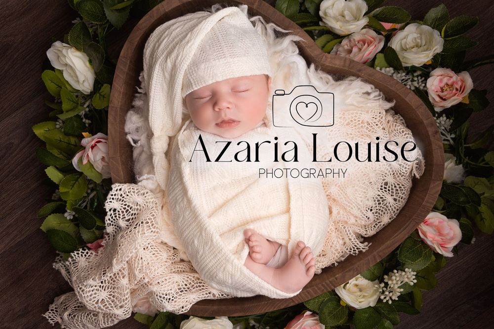 A sleeping newborn wrapped in white lace is nestled in a heart-shaped basket surrounded by pink roses.