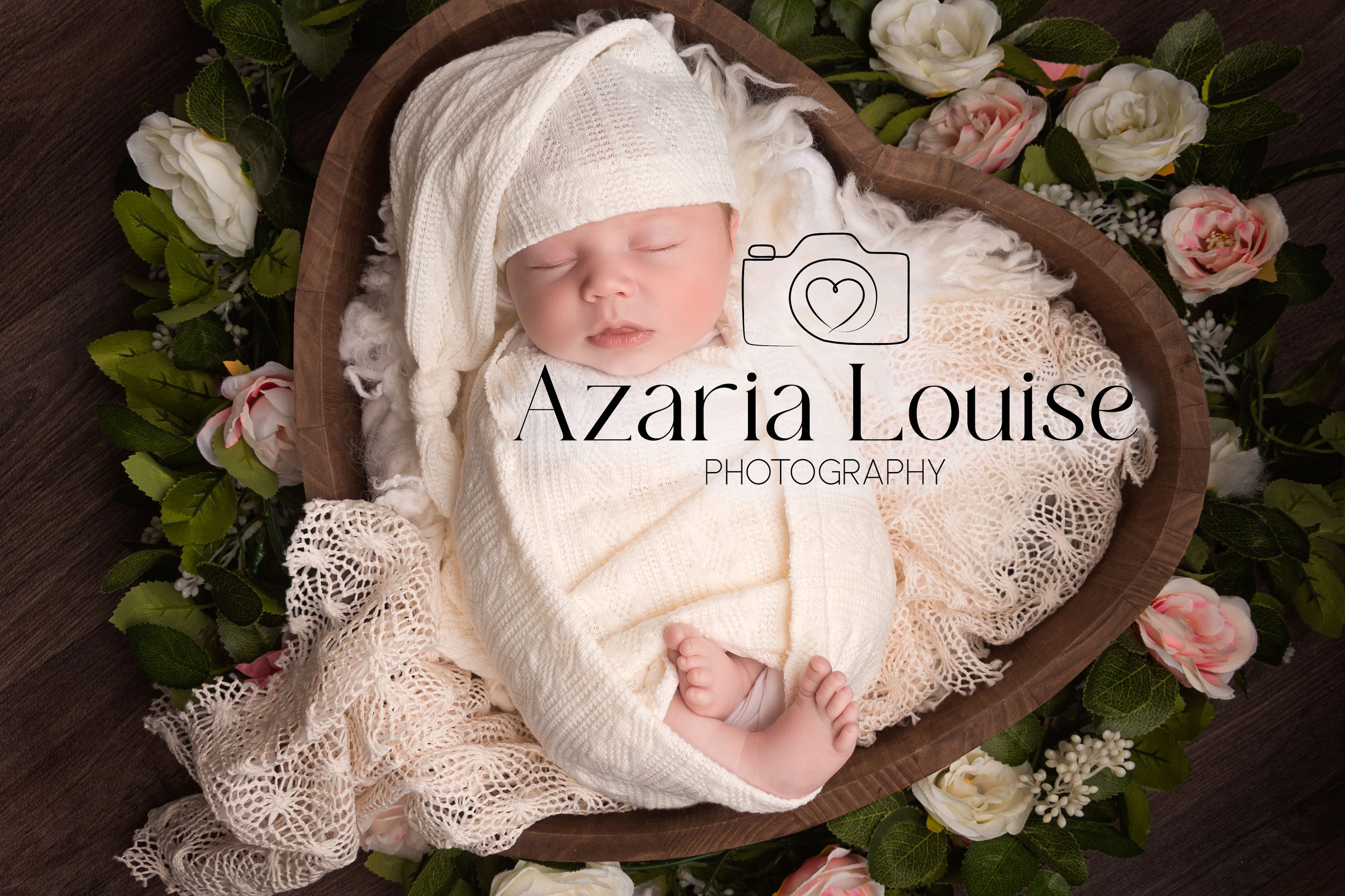 A sleeping newborn wrapped in white lace is nestled in a heart-shaped basket surrounded by pink roses.