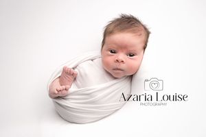 A swaddled newborn baby lies on a white backdrop with a photographer's logo in the corner.