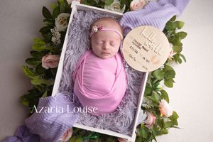 A newborn swaddled in pink fabric rests in a gray crate surrounded by greenery and roses with a wooden name tag.