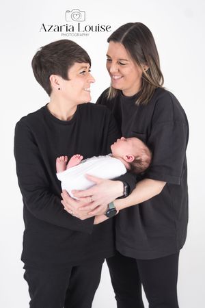A couple wearing matching black outfits holds a newborn baby in white against a light studio backdrop.