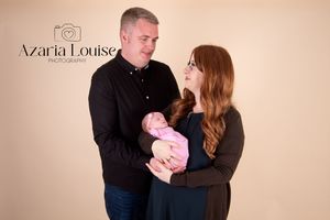 A couple in dark clothing gaze lovingly at their newborn baby wrapped in pink against a neutral studio backdrop.