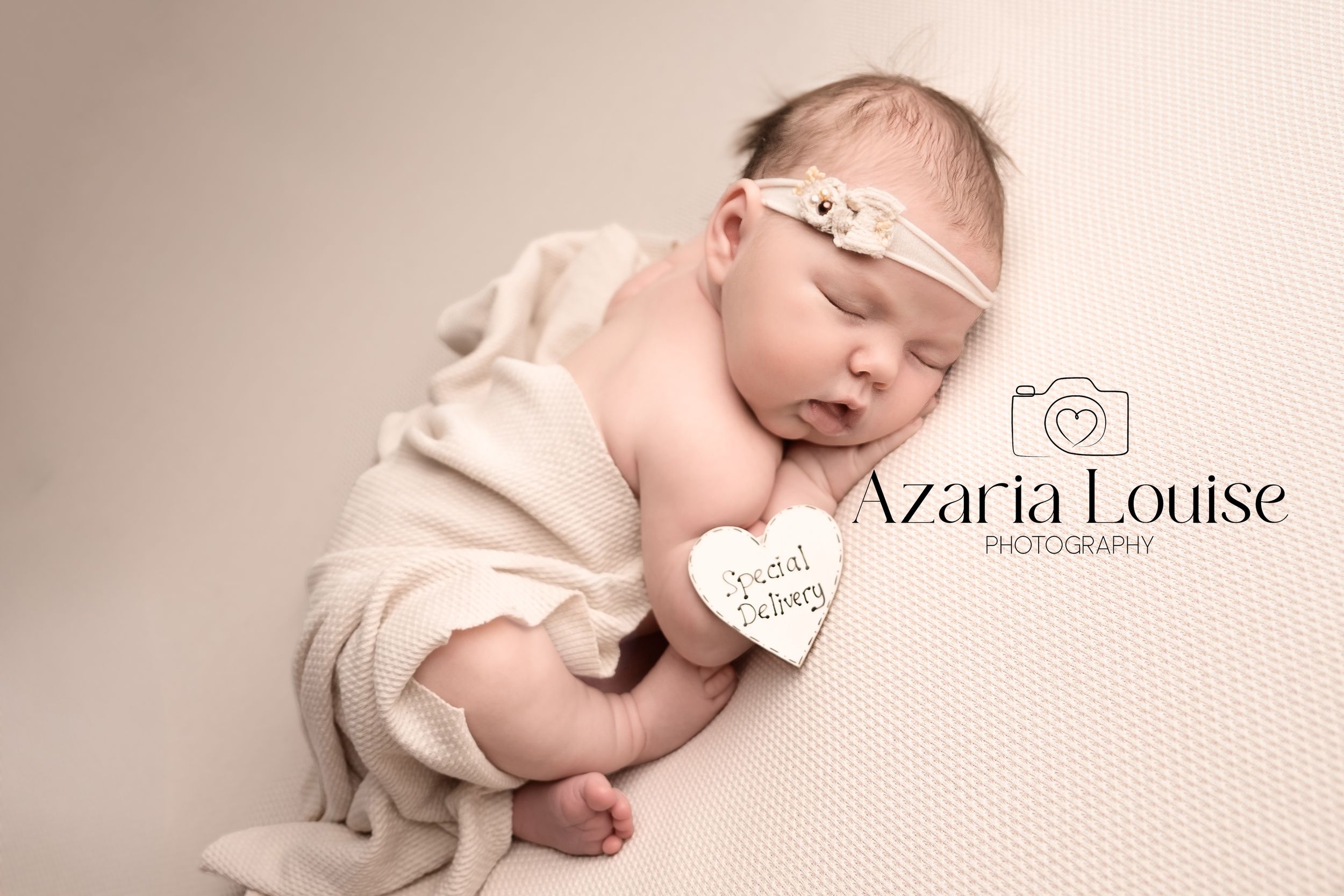 Newborn baby sleeping peacefully on beige backdrop with delicate headband and heart tag from Azaria Louise Photography.